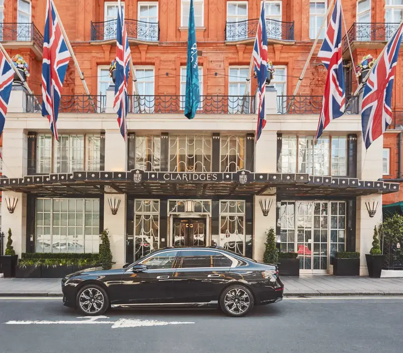 A sleek black luxury car parked outside the entrance of Claridge’s hotel in London, with Union Jack and teal flags hanging above the grand Art Deco façade of red brick and cream stone.