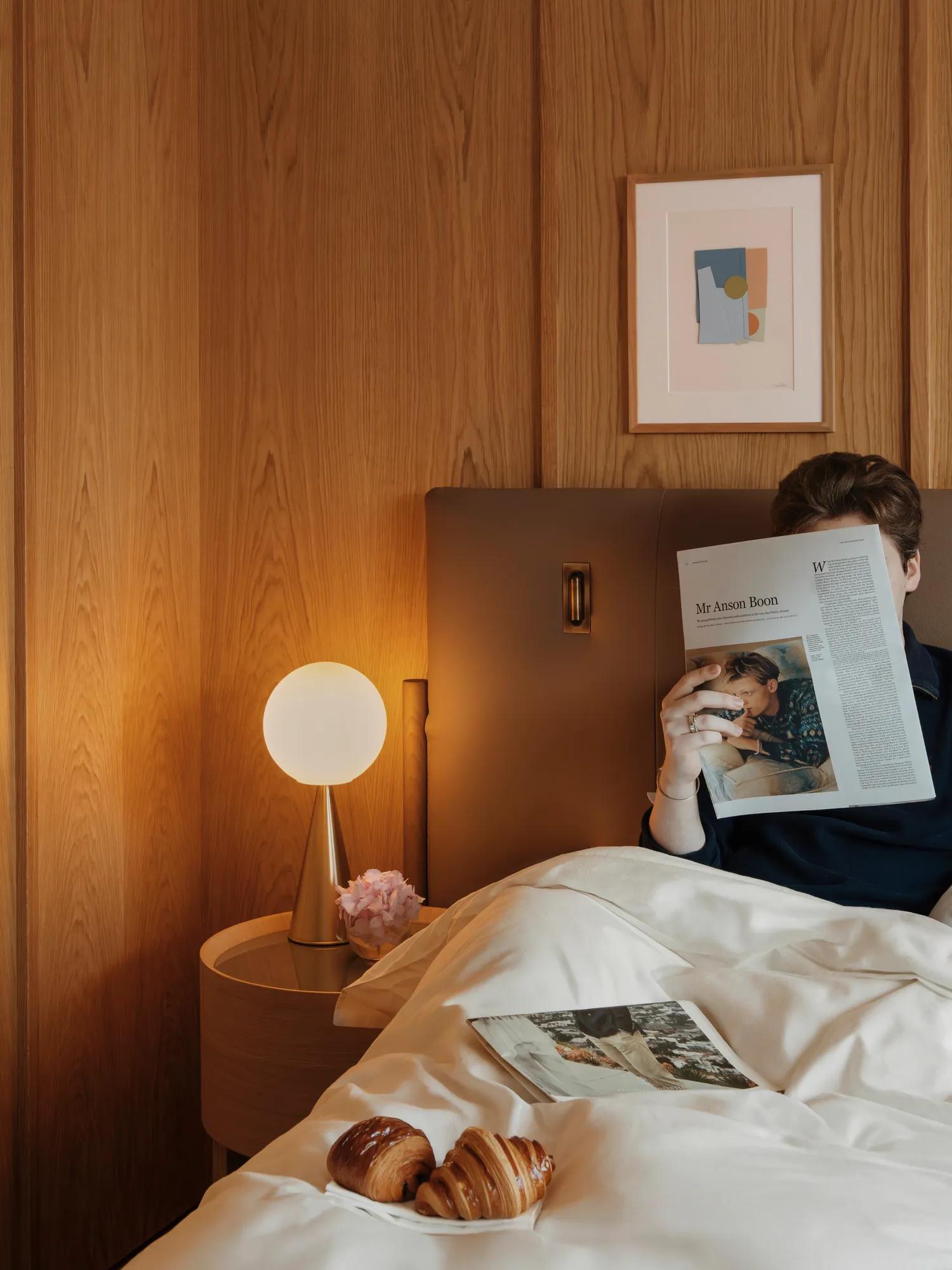 Person reading a newspaper in bed with a bedside lamp, pastries and a magazine on white linens