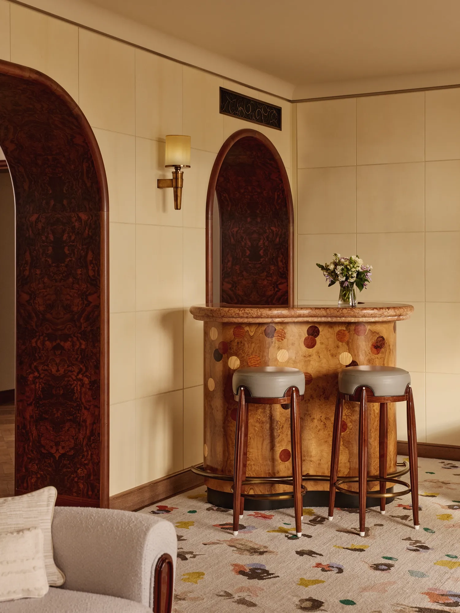 Stylish in-room bar with a curved wooden counter featuring circular inlays, two upholstered bar stools, and arched doorways with dark wood paneling, set against soft beige walls.
