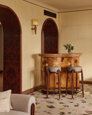 Stylish in-room bar with a curved wooden counter featuring circular inlays, two upholstered bar stools, and arched doorways with dark wood paneling, set against soft beige walls.