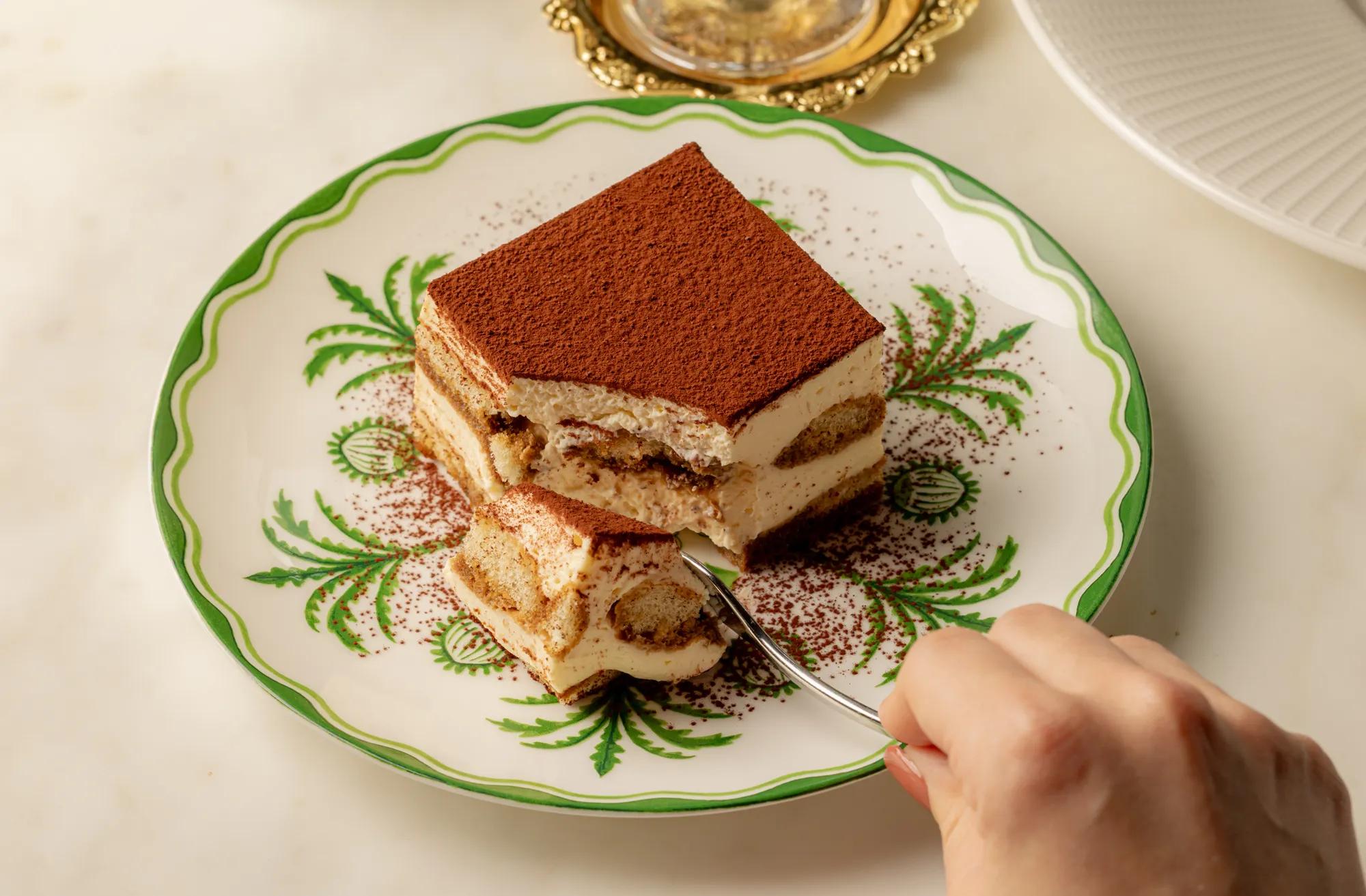 Hand holding a spoonful of tiramisu above a cocoa-dusted slice, served on a decorative green-patterned plate.