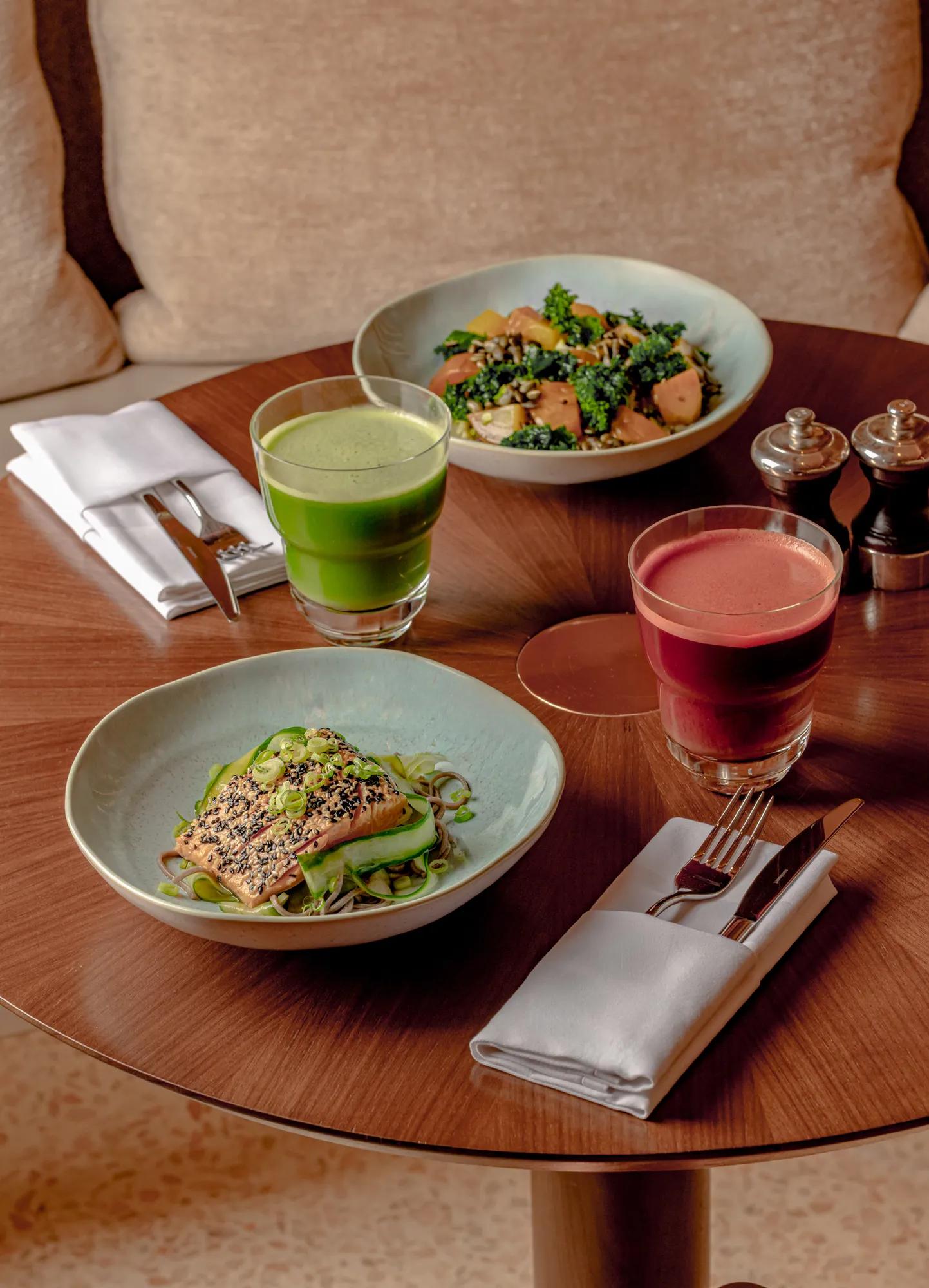 A table set with a healthy meal of grilled salmon topped with sesame seeds and green onions on noodles, alongside two vibrant drinks - green juice and beetroot juice - on a wooden table with utensils and napkins.