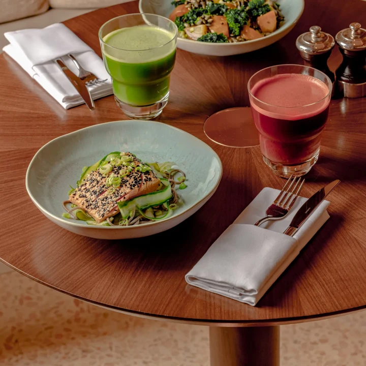 A table set with a healthy meal of grilled salmon topped with sesame seeds and green onions on noodles, alongside two vibrant drinks - green juice and beetroot juice - on a wooden table with utensils and napkins.