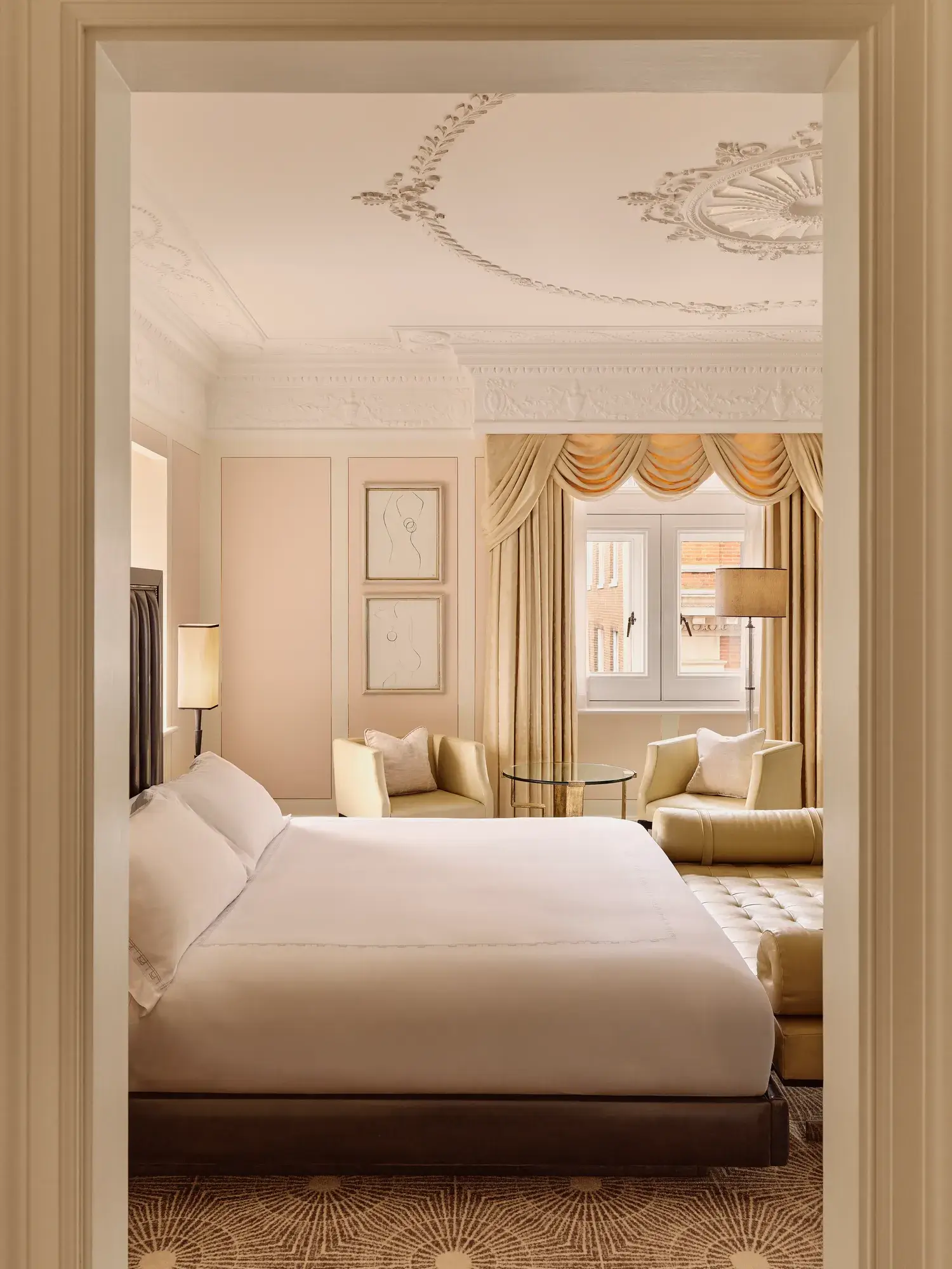 Elegant bedroom with ornate ceiling, draped window, cream armchairs and upholstered bed.