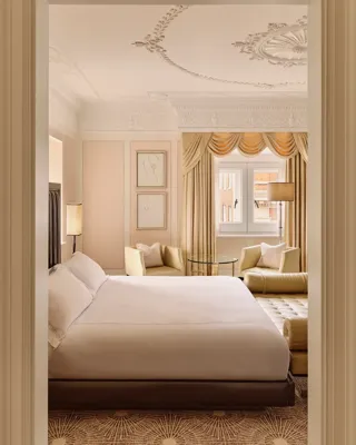 Elegant bedroom with ornate ceiling, draped window, cream armchairs and upholstered bed.