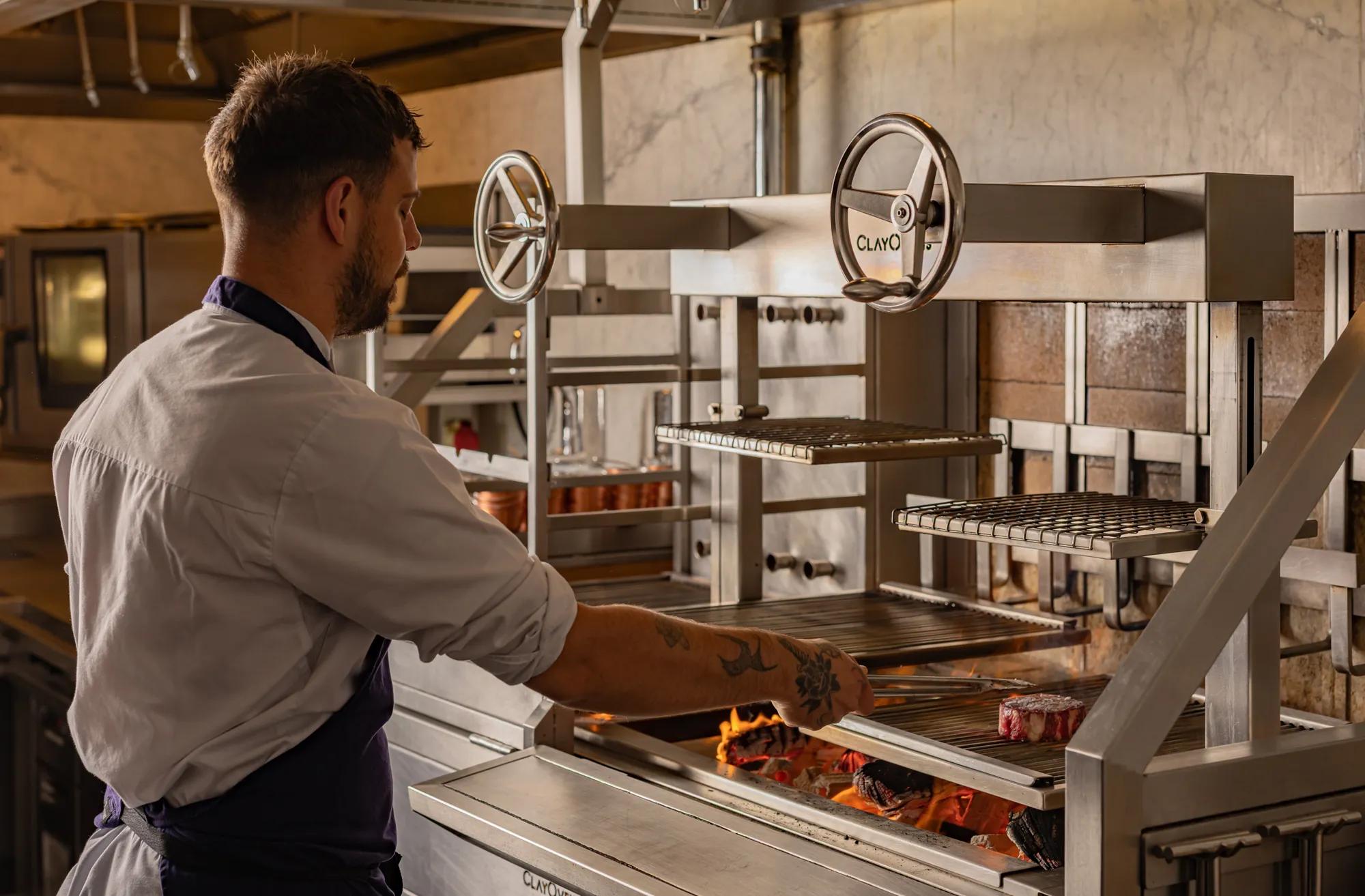 **Alt text (WCAG 2.2 AA, under 125 characters):**

Chef grills steaks over an open flame in a professional kitchen using an adjustable charcoal grill.
