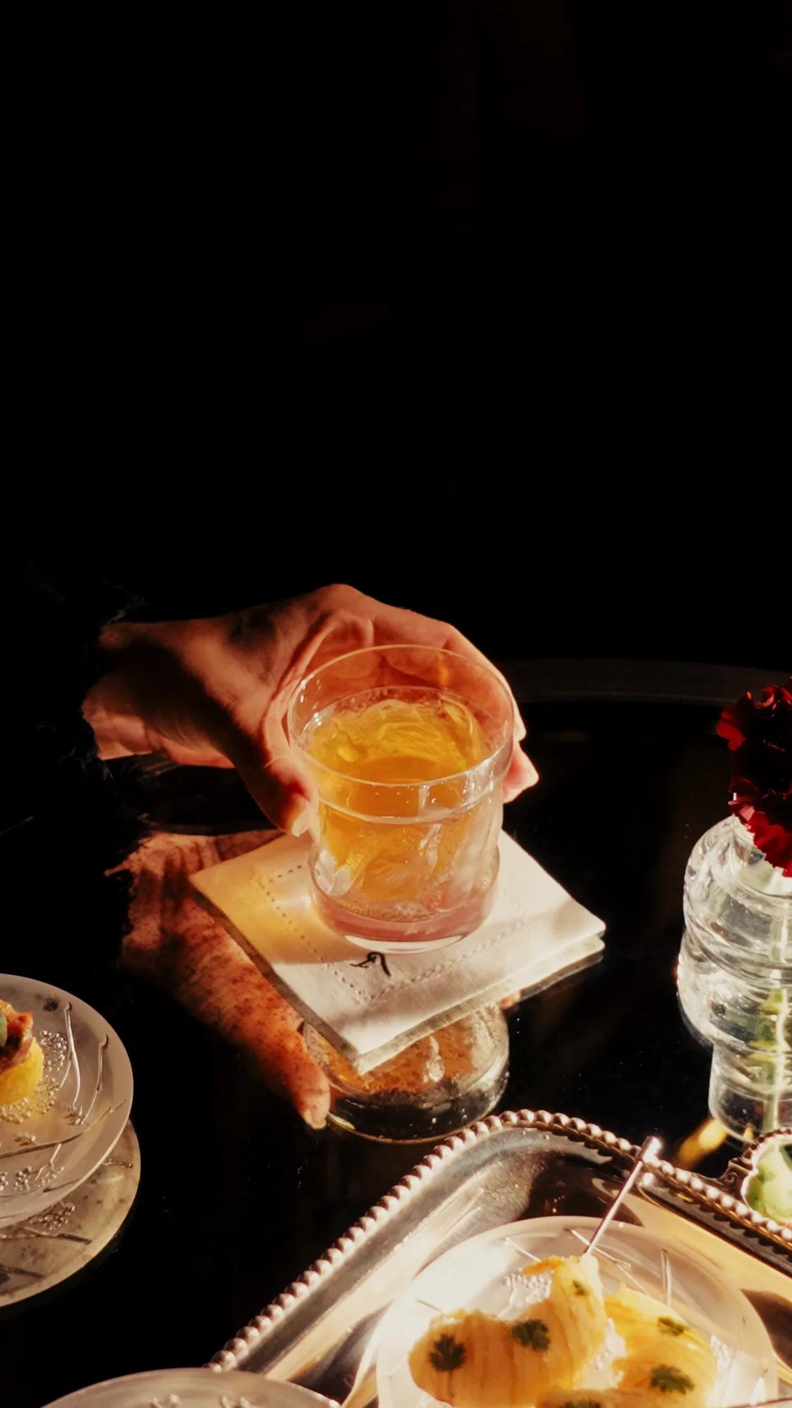 A hand holding an amber cocktail above a softly lit table, surrounded by elegant bar bites and glassware in an intimate, atmospheric setting.