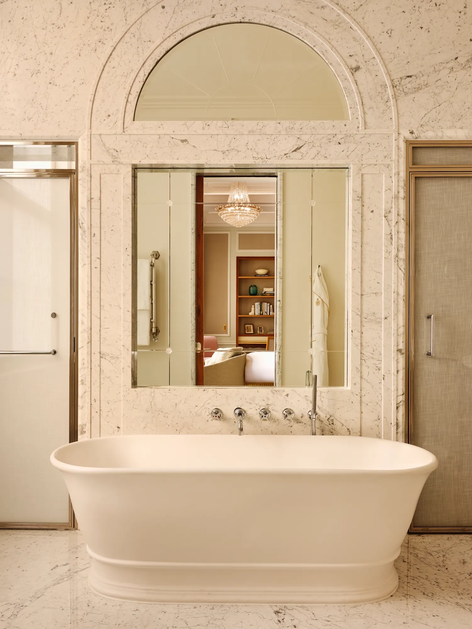 Luxurious marble bathroom with a freestanding white bathtub, large mirror reflecting a chandelier and bedroom beyond, and frosted glass doors on either side.