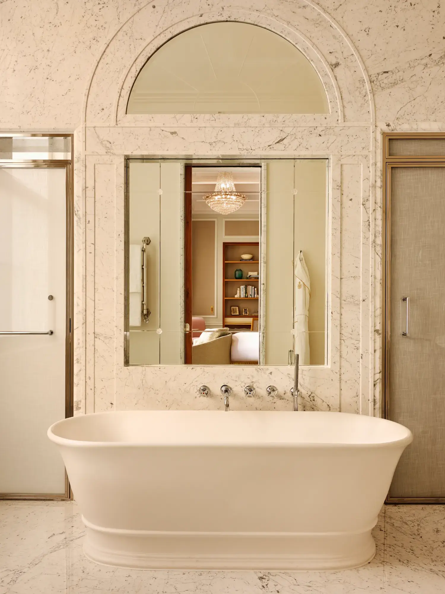 Luxurious marble bathroom with a freestanding white bathtub, large mirror reflecting a chandelier and bedroom beyond, and frosted glass doors on either side.