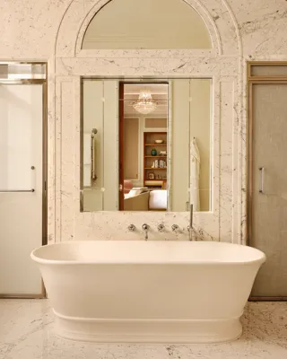Luxurious marble bathroom with a freestanding white bathtub, large mirror reflecting a chandelier and bedroom beyond, and frosted glass doors on either side.