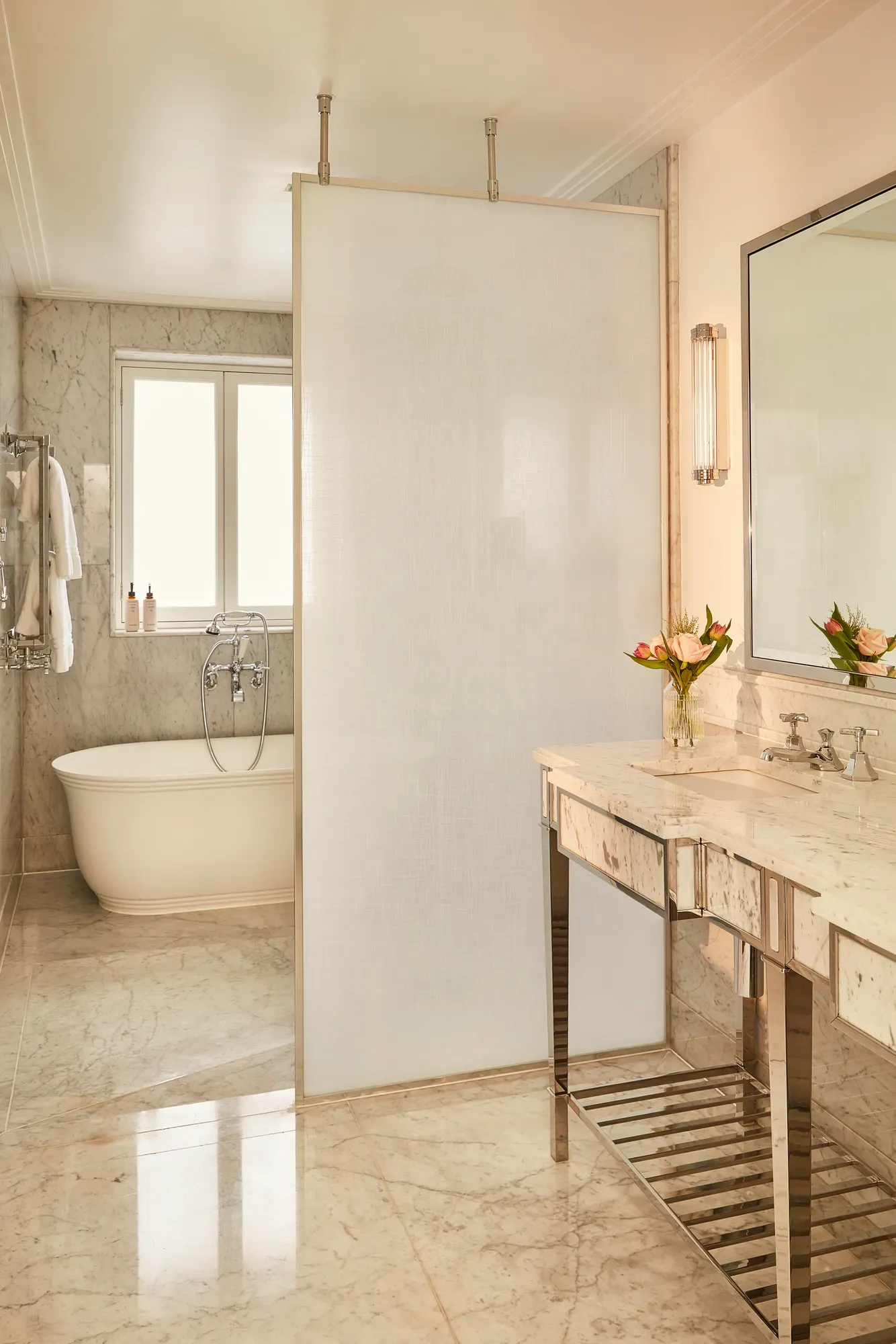 Elegant marble bathroom with a freestanding bathtub, large vanity with flowers, and a frosted glass divider separating the spaces.