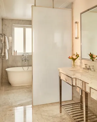 Elegant marble bathroom with a freestanding bathtub, large vanity with flowers, and a frosted glass divider separating the spaces.