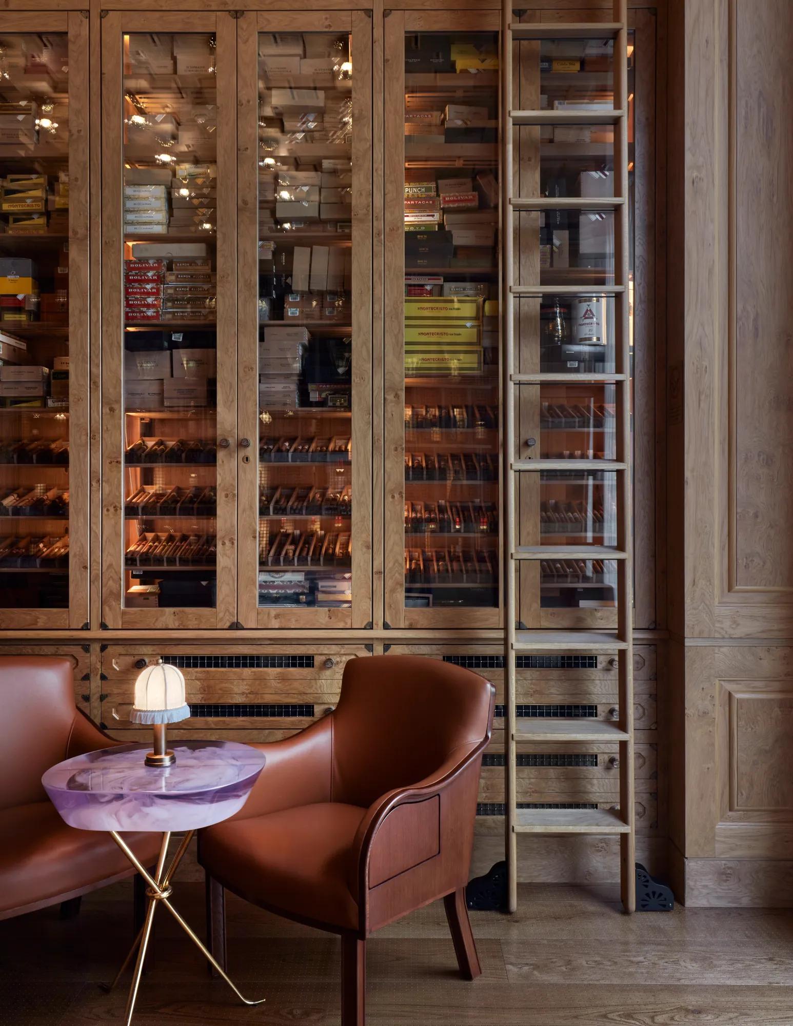 Warm wood-panelled cigar room with glass cabinets of cigars, a sliding ladder, and two leather chairs around a small purple table.