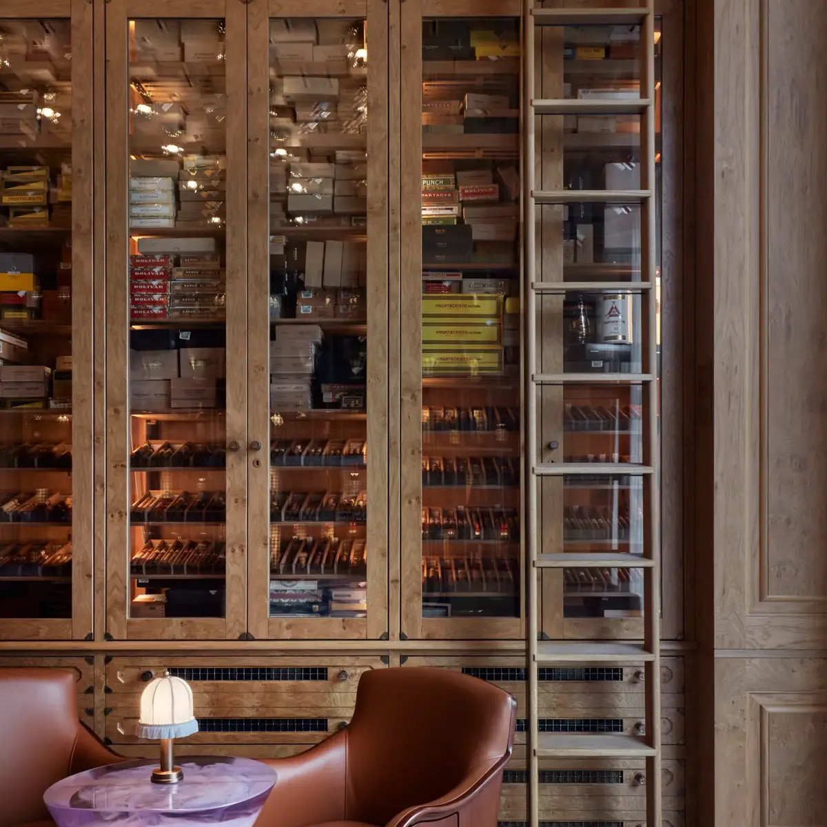 Warm wood-panelled cigar room with glass cabinets of cigars, a sliding ladder, and two leather chairs around a small purple table.