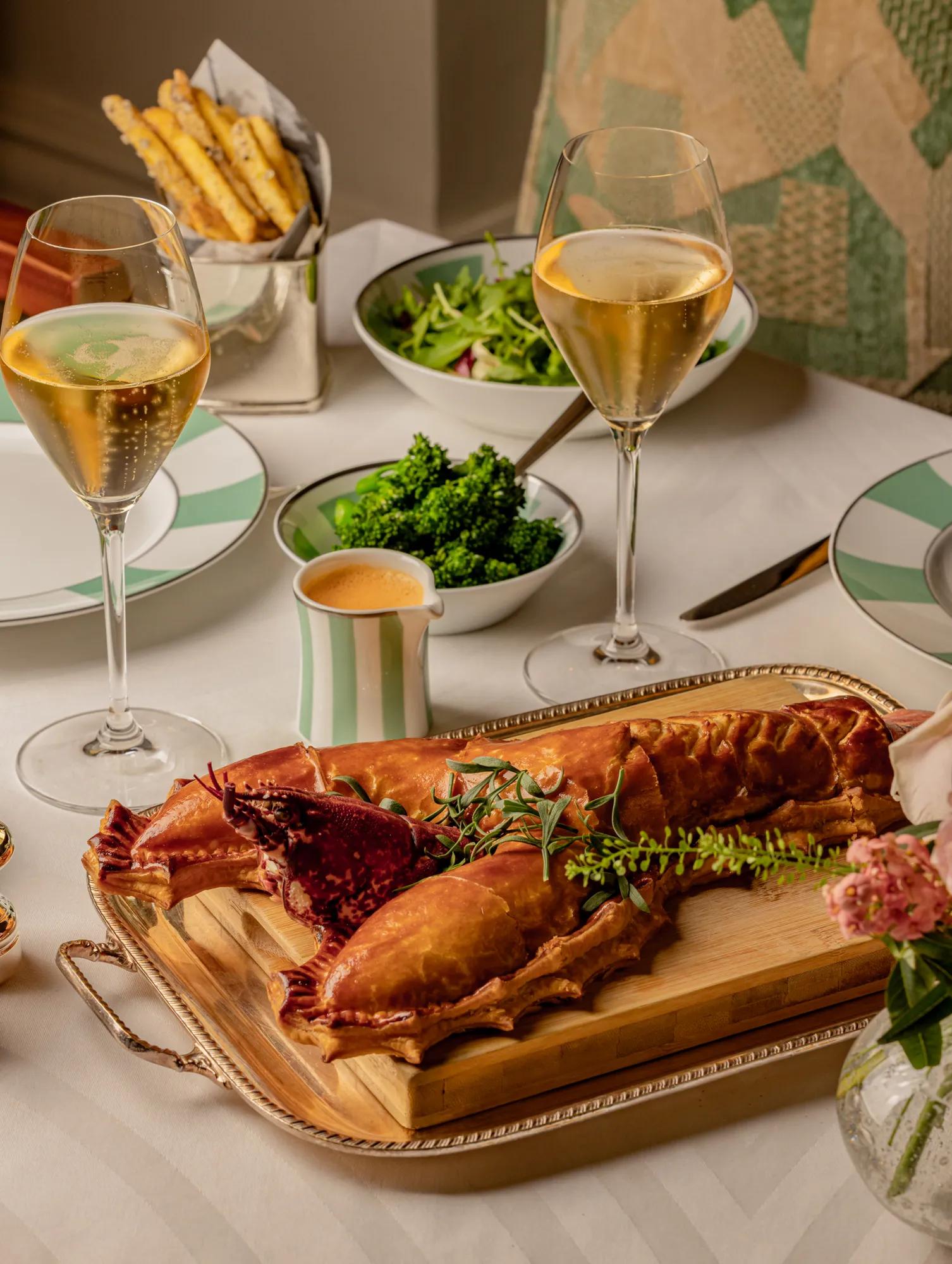 Lobster dish served on a wooden board with champagne, salad, and sides arranged on a neatly set dining table.