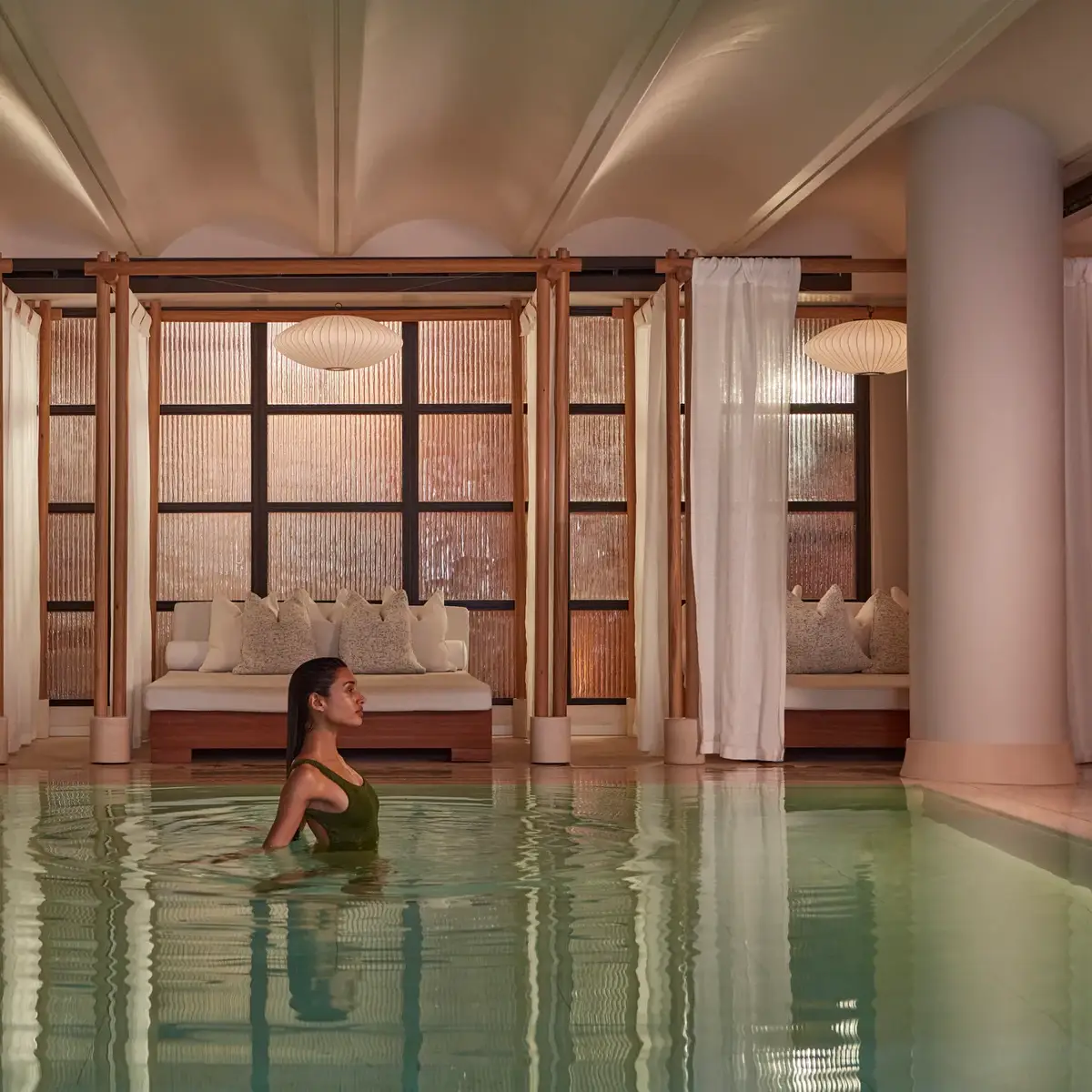 A woman wades in a calm indoor pool, with curtained lounge beds and soft warm lighting behind her.