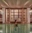 A woman wades in a calm indoor pool, with curtained lounge beds and soft warm lighting behind her.