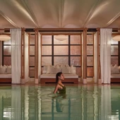 A woman wades in a calm indoor pool, with curtained lounge beds and soft warm lighting behind her.