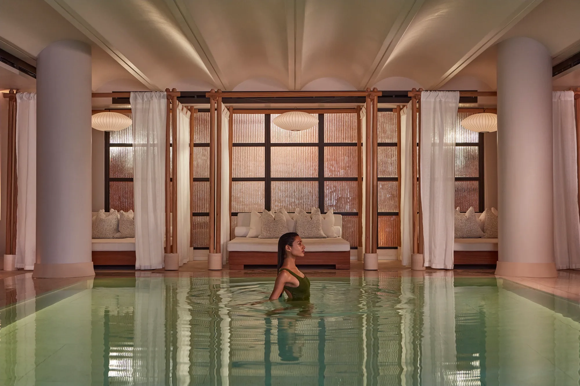 A woman wades in a calm indoor pool, with curtained lounge beds and soft warm lighting behind her.