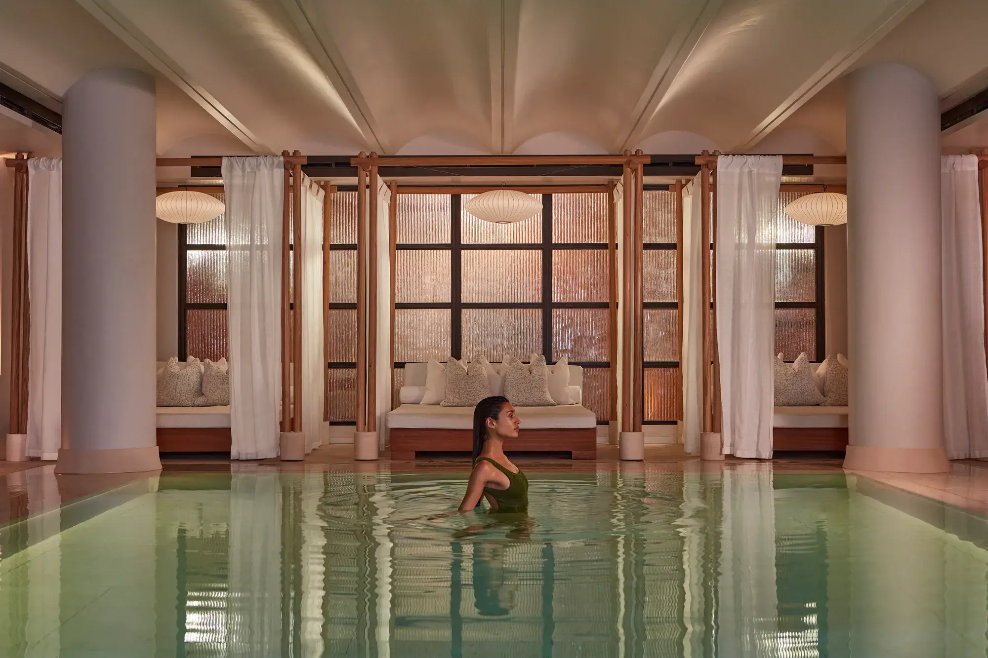 A woman wades in a calm indoor pool, with curtained lounge beds and soft warm lighting behind her.