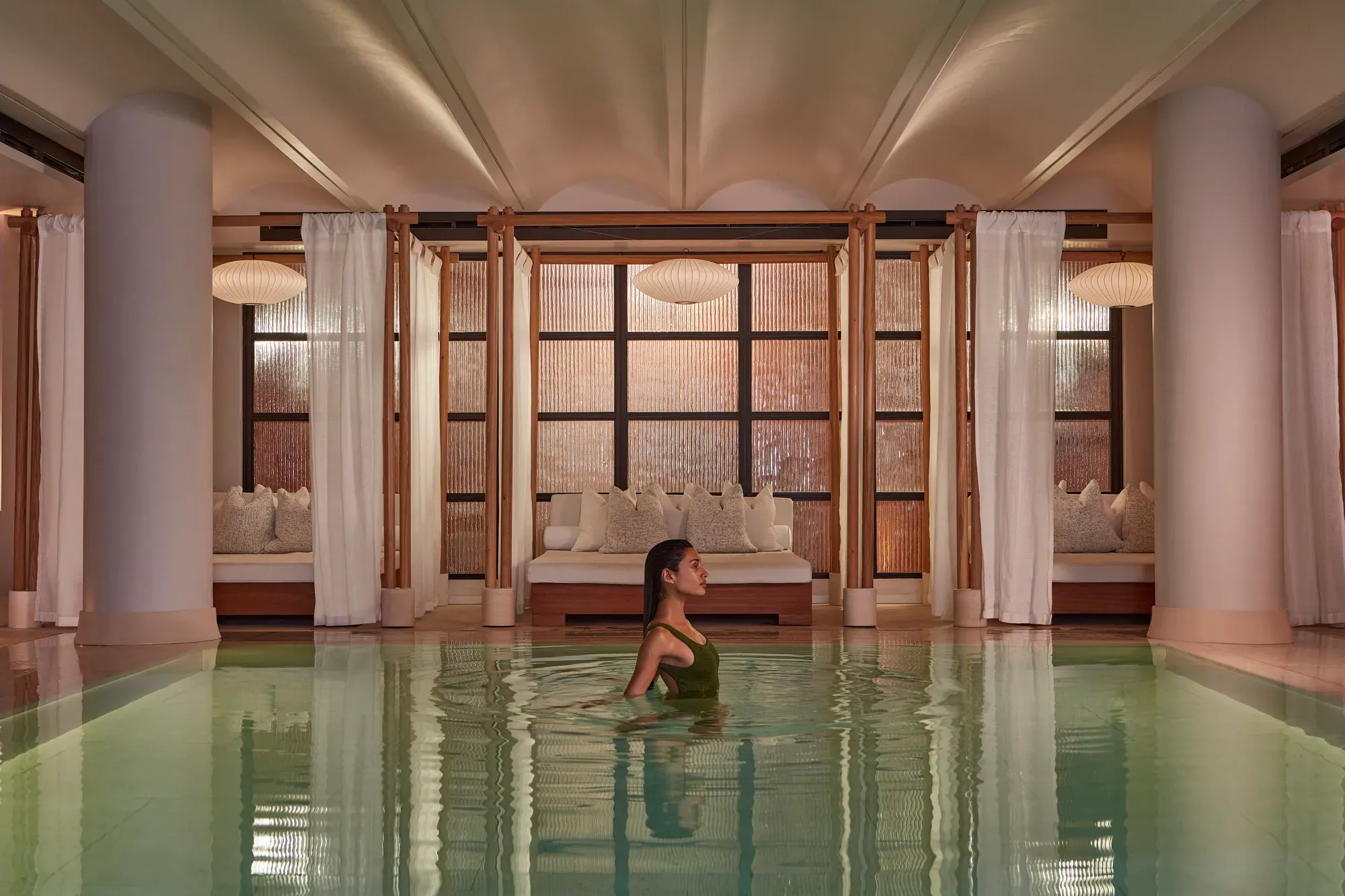 A woman wades in a calm indoor pool, with curtained lounge beds and soft warm lighting behind her.
