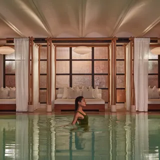 A woman wades in a calm indoor pool, with curtained lounge beds and soft warm lighting behind her.