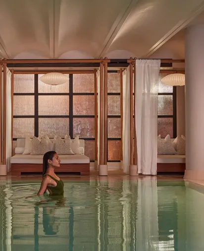 A woman wades in a calm indoor pool, with curtained lounge beds and soft warm lighting behind her.