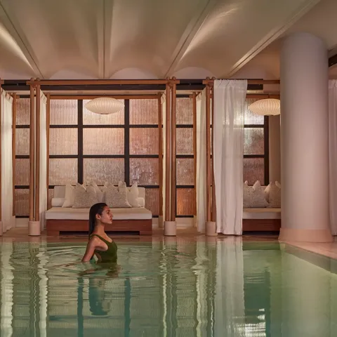 A woman wades in a calm indoor pool, with curtained lounge beds and soft warm lighting behind her.