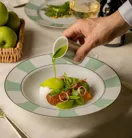 Hand pouring green sauce over plated starter with vegetables, beside wine glass and bottle.