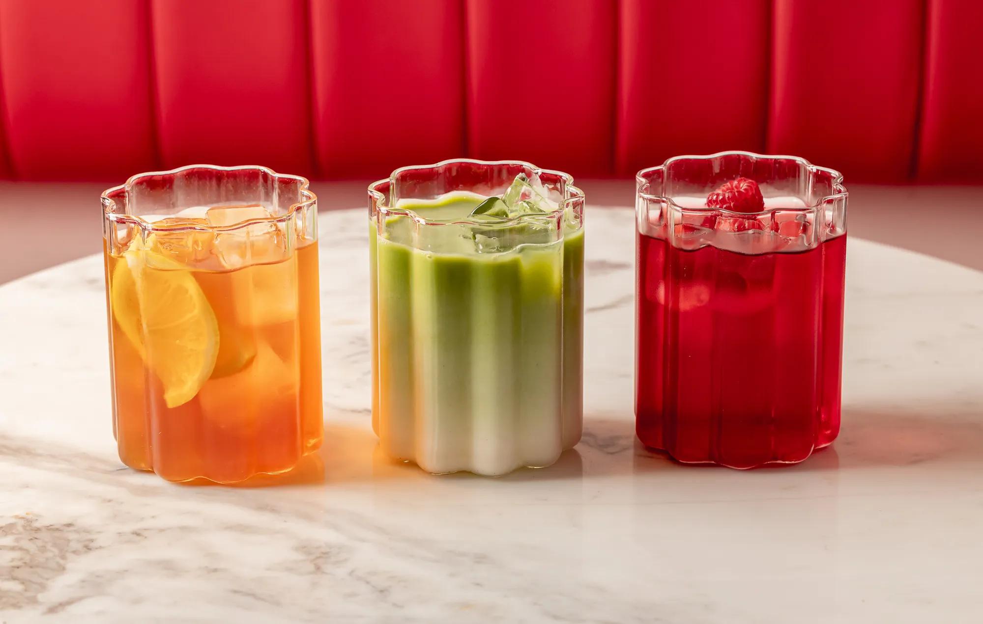 Three colourful iced drinks—citrus, green matcha, and raspberry—on a marble table against a red banquette.