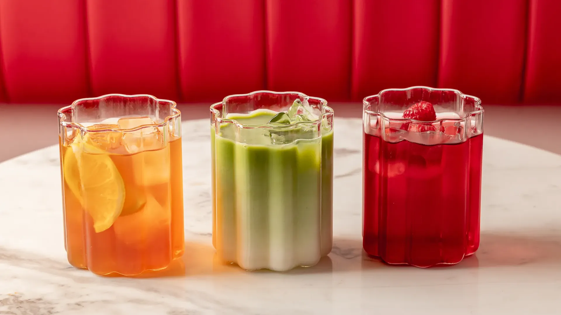 Three colourful iced drinks—citrus, green matcha, and raspberry—on a marble table against a red banquette.