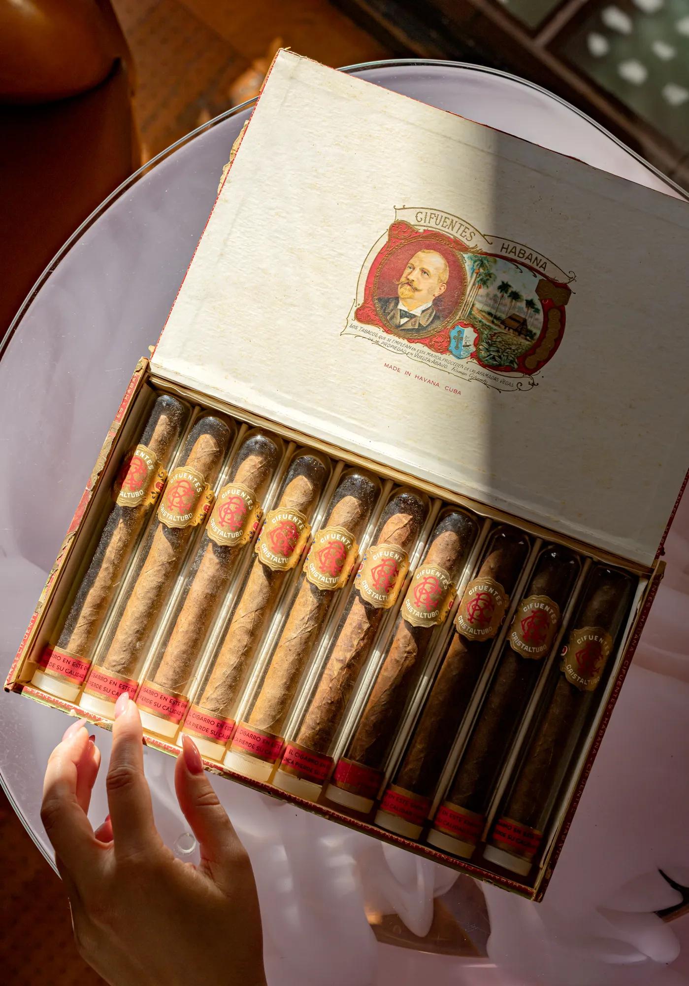 Open cigar box with neatly arranged cigars, a hand reaching toward them on a round table