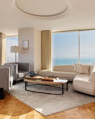 Salon moderne avec canapé blanc incurvé, fauteuils gris et table basse, doté de baies vitrées offrant une vaste vue sur la mer.