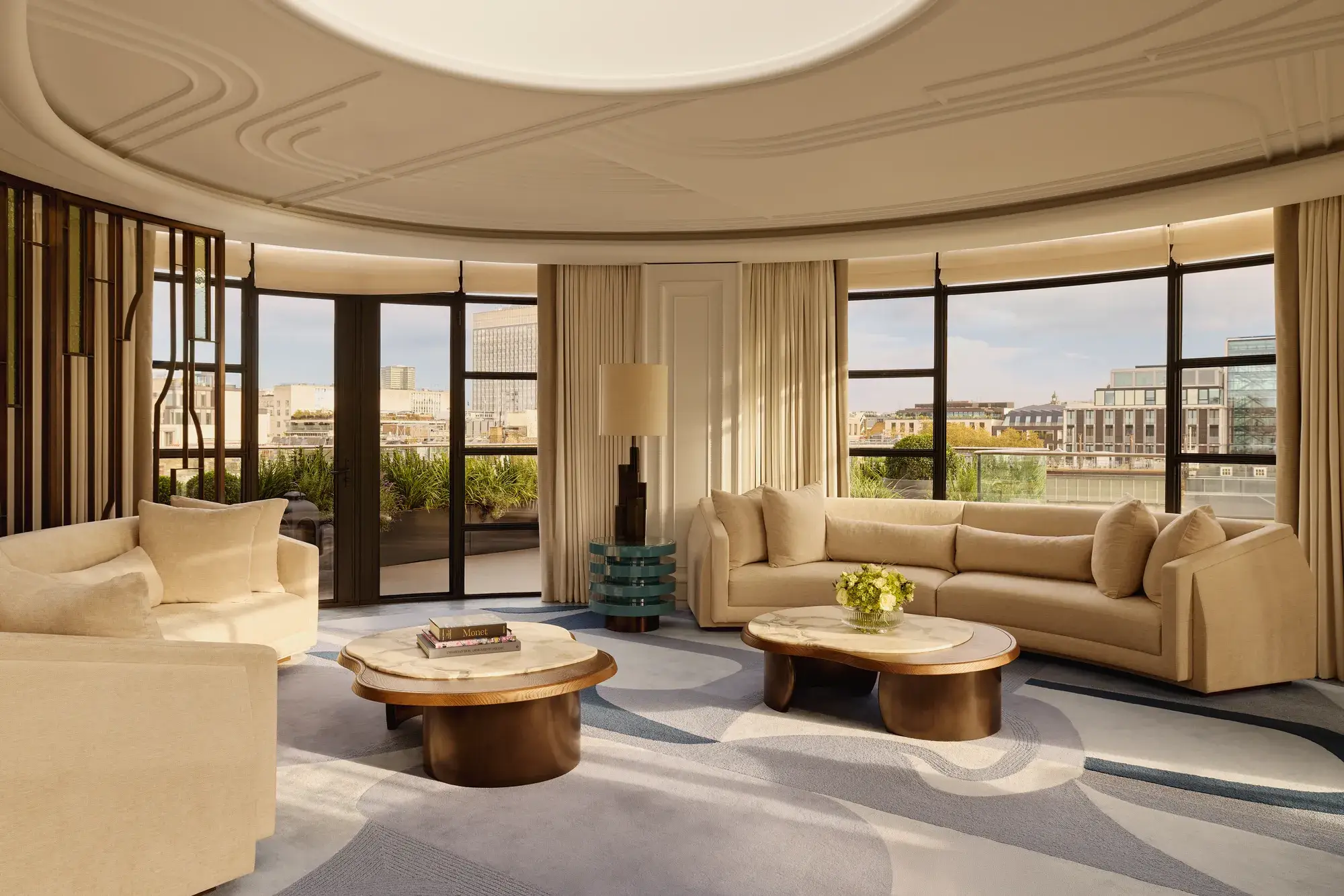 Spacious curved living room with large windows offering city views, featuring cream sofas, marble-topped coffee tables, a patterned blue and beige rug, and soft natural light filtering through full-length curtains.