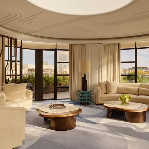 Spacious curved living room with large windows offering city views, featuring cream sofas, marble-topped coffee tables, a patterned blue and beige rug, and soft natural light filtering through full-length curtains.