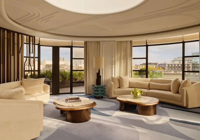 Spacious curved living room with large windows offering city views, featuring cream sofas, marble-topped coffee tables, a patterned blue and beige rug, and soft natural light filtering through full-length curtains.