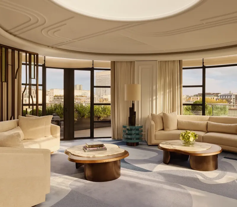 Spacious curved living room with large windows offering city views, featuring cream sofas, marble-topped coffee tables, a patterned blue and beige rug, and soft natural light filtering through full-length curtains.