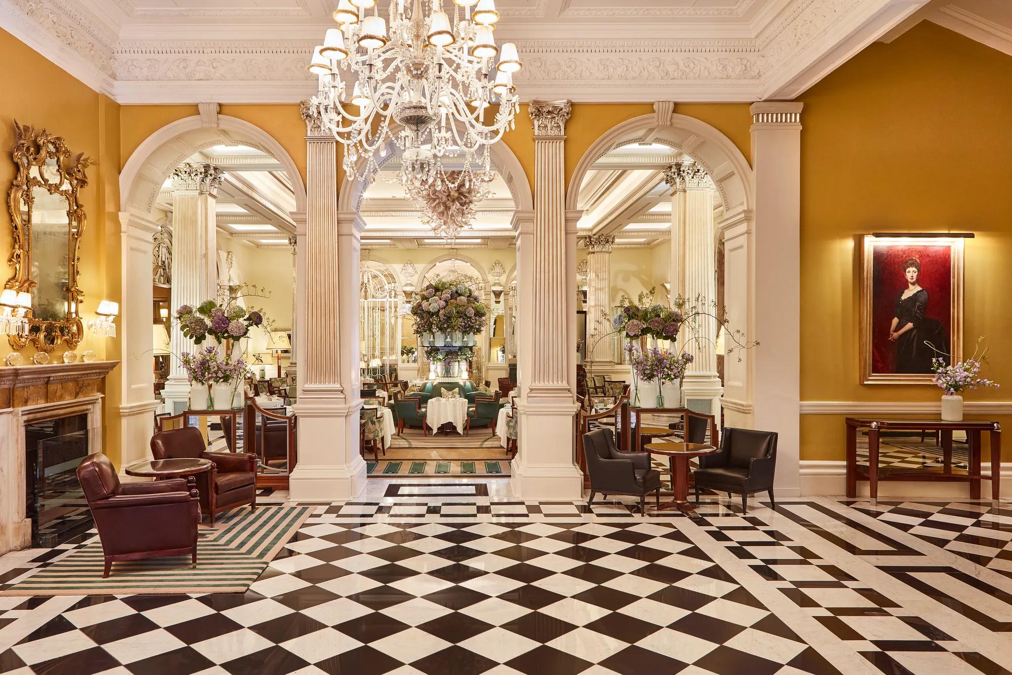 An elegant hotel lobby with a black-and-white chequered marble floor, crystal chandeliers, and classical columns leading into a refined lounge area decorated with large floral arrangements and comfortable seating.