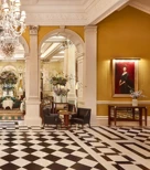 An elegant hotel lobby with a black-and-white chequered marble floor, crystal chandeliers, and classical columns leading into a refined lounge area decorated with large floral arrangements and comfortable seating.