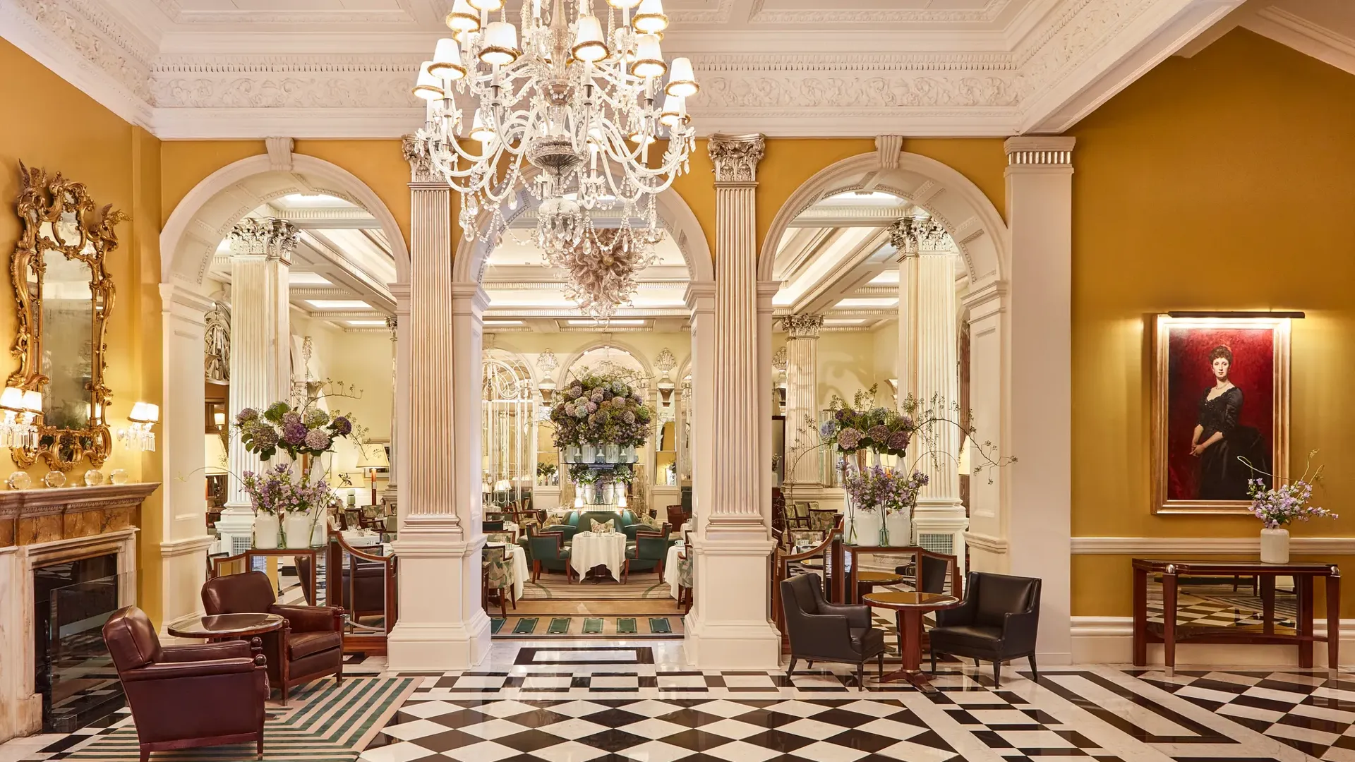 An elegant hotel lobby with a black-and-white chequered marble floor, crystal chandeliers, and classical columns leading into a refined lounge area decorated with large floral arrangements and comfortable seating.