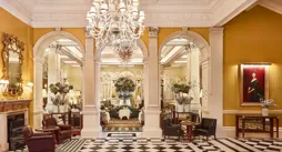 An elegant hotel lobby with a black-and-white chequered marble floor, crystal chandeliers, and classical columns leading into a refined lounge area decorated with large floral arrangements and comfortable seating.