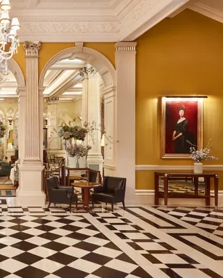 An elegant hotel lobby with a black-and-white chequered marble floor, crystal chandeliers, and classical columns leading into a refined lounge area decorated with large floral arrangements and comfortable seating.