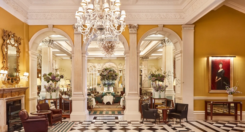 An elegant hotel lobby with a black-and-white chequered marble floor, crystal chandeliers, and classical columns leading into a refined lounge area decorated with large floral arrangements and comfortable seating.