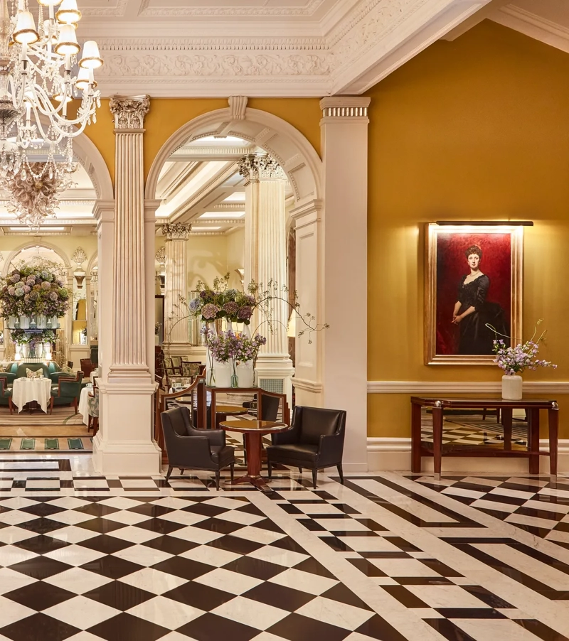 An elegant hotel lobby with a black-and-white chequered marble floor, crystal chandeliers, and classical columns leading into a refined lounge area decorated with large floral arrangements and comfortable seating.