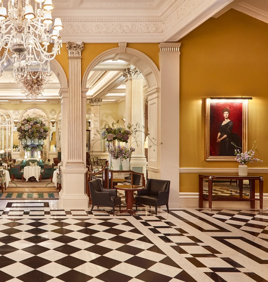 An elegant hotel lobby with a black-and-white chequered marble floor, crystal chandeliers, and classical columns leading into a refined lounge area decorated with large floral arrangements and comfortable seating.