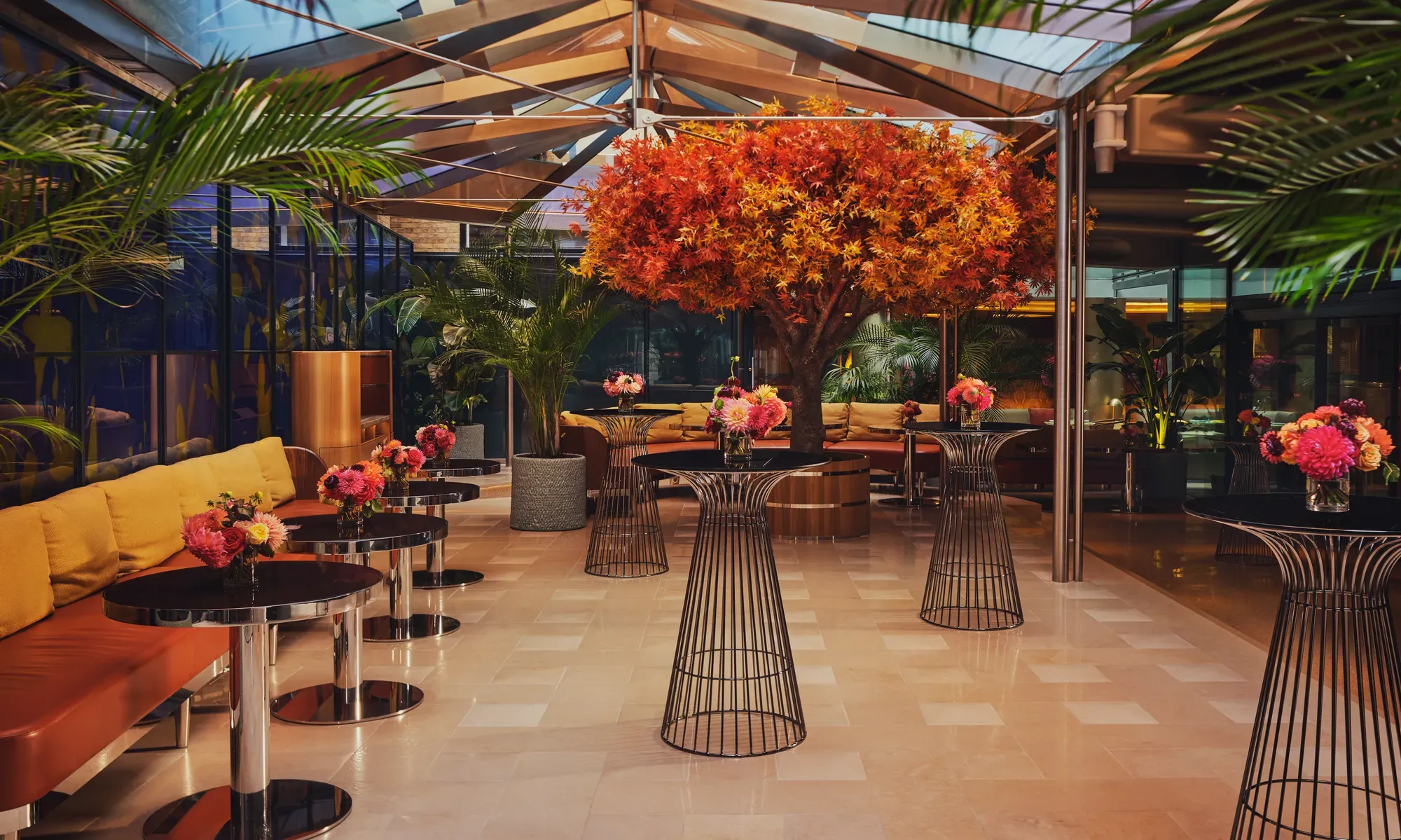 Glass-roofed courtyard with central autumn-hued tree, cocktail tables, vibrant floral arrangements, and plush seating surrounded by greenery.