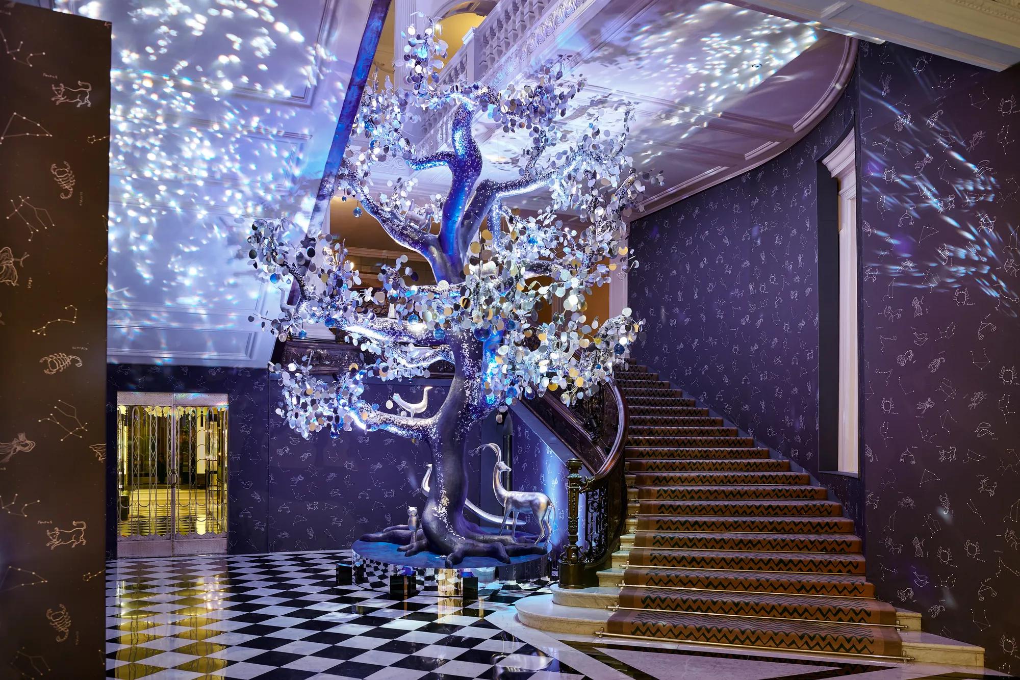 Grand hotel lobby with sweeping staircase and checkered floor, featuring illuminated sculptural tree casting star-like reflections against constellation-patterned walls.