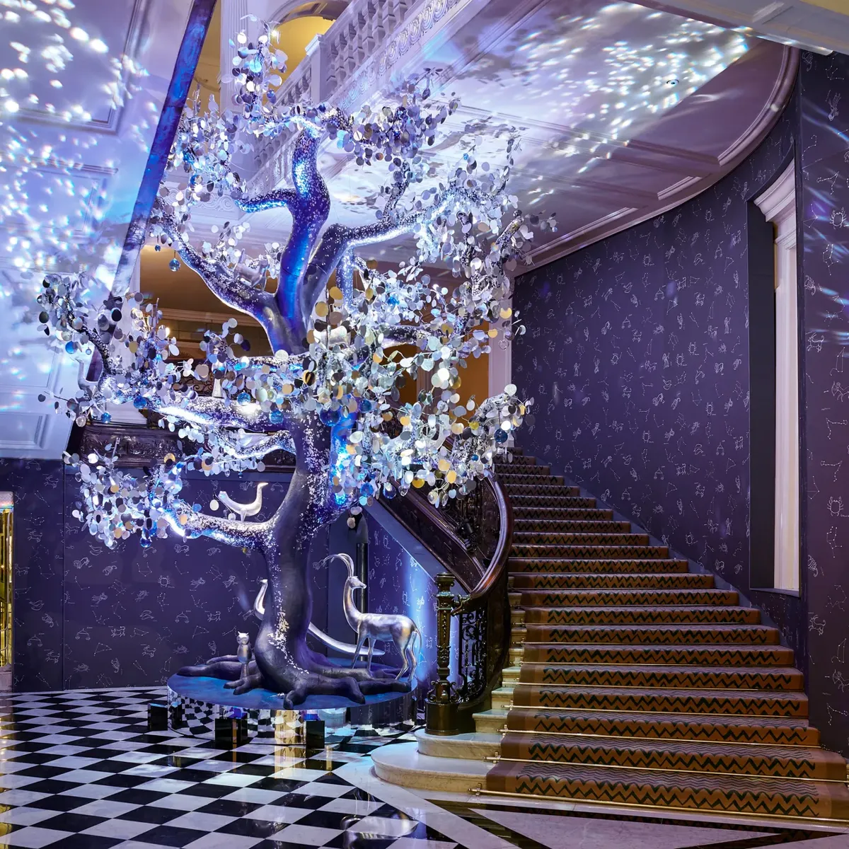 Grand hotel lobby with sweeping staircase and checkered floor, featuring illuminated sculptural tree casting star-like reflections against constellation-patterned walls.