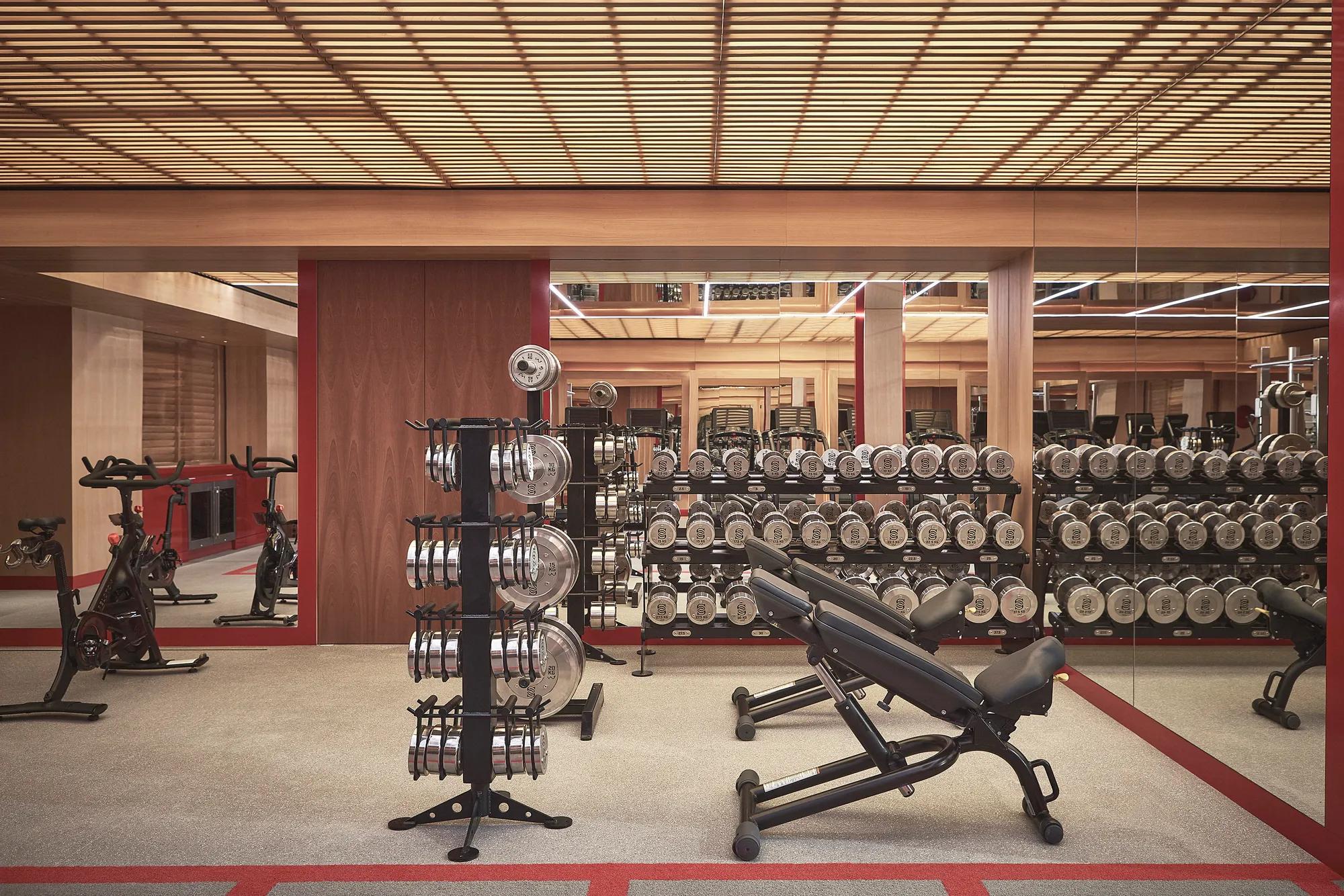 Modern gym with free weights, adjustable benches, mirrored walls, and exercise bikes under warm wood-paneled ceilings.