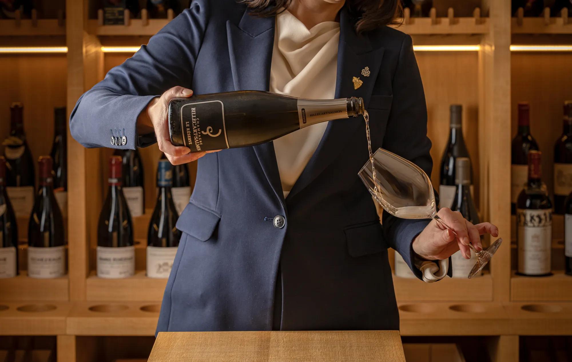 A bottle of sparkling wine is poured into a wine glass in front of a softly lit wine display lined with bottles.